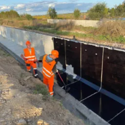 Dw&oacute;ch pracownik&oacute;w w pomarańczowych kombinezonach zabezpiecza betonowe koryto, malując je czarną substancją za pomocą wałka na długim uchwycie. W tle widoczne pole i niebo z chmurami.