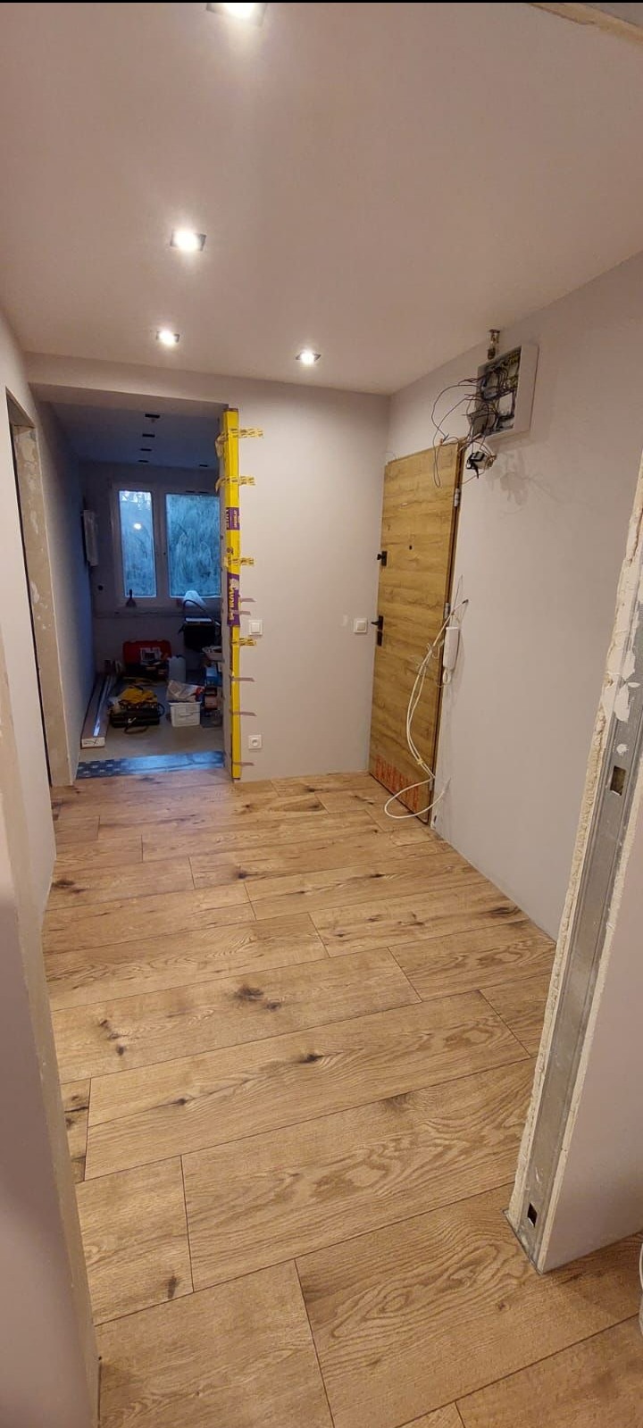 Wnętrze w trakcie remontu: Ułożona podłoga z paneli imitujących drewno, widoczna ościeżnica, niezabezpieczona instalacja elektryczna i poziomica na ścianie przy wejściu do kolejnego pomieszczenia.