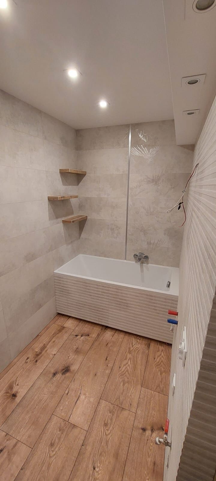 Wykończona łazienka w Kielcach z drewnianą podłogą, płytkami w odcieniach beżu na ścianach, białą wanną z poziomymi żłobieniami i trzema drewnianymi półkami.
