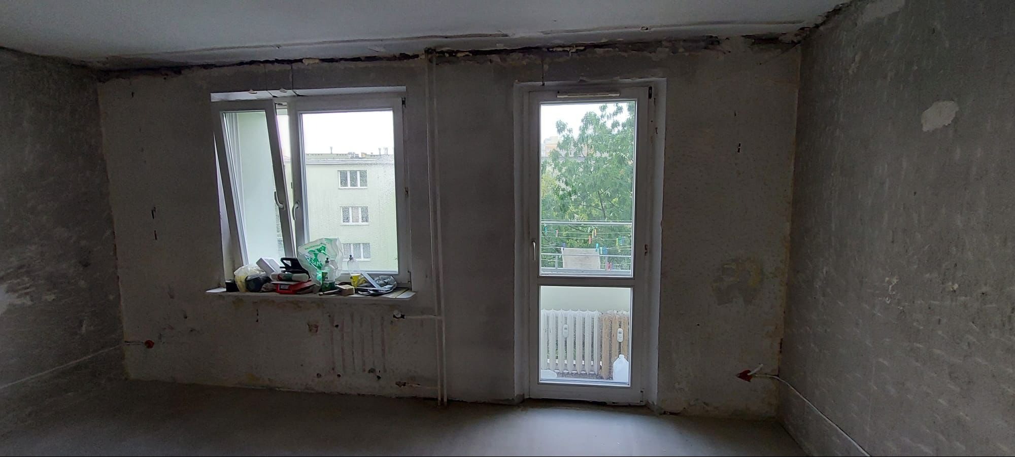 Pokój w trakcie remontu z surowymi ścianami, widoczne okno z parapetem zastawionym narzędziami i materiałami budowlanymi, oraz balkon z widokiem na zieleń.