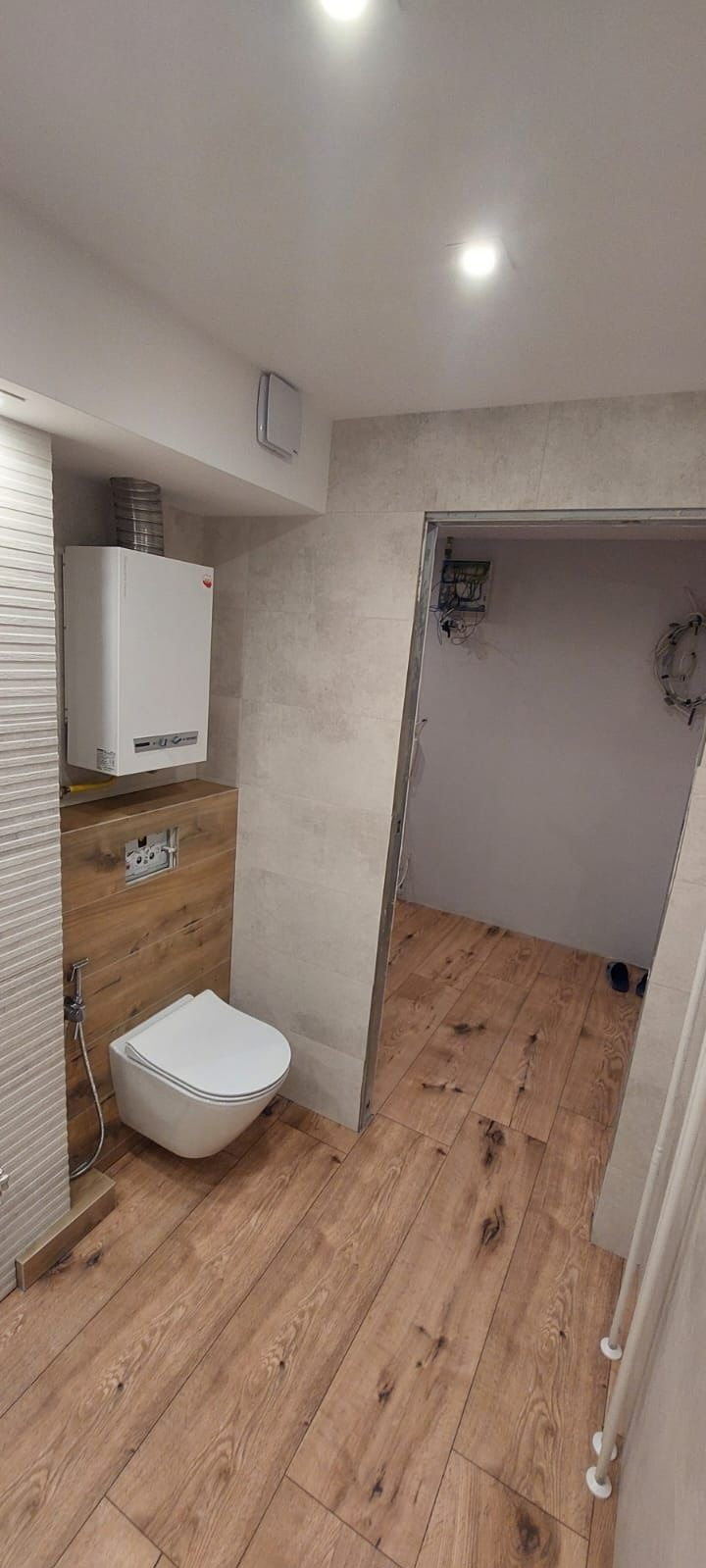 Nowoczesna łazienka z wiszącą toaletą, grzejnikiem i drewnopodobną podłogą, widok na sąsiednie pomieszczenie z niedokończonymi pracami elektrycznymi.