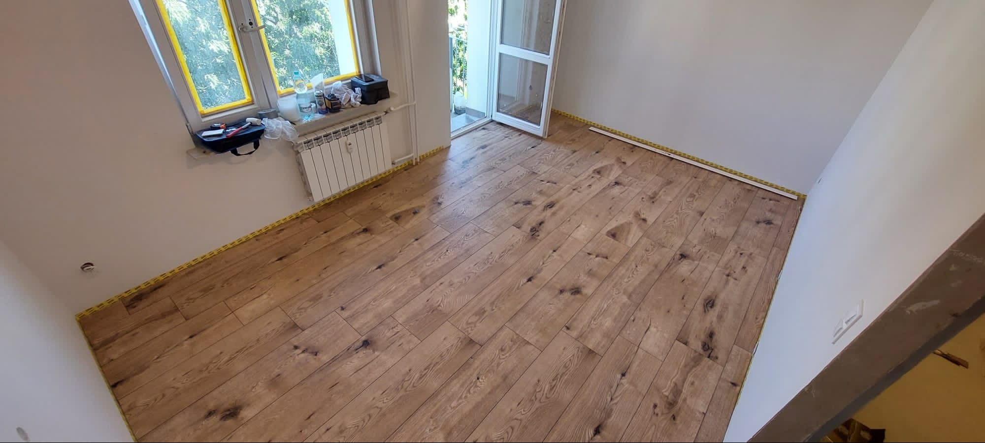 Nowo ułożony parkiet o wzorze rustykalnym z widocznymi sękami, zabezpieczony taśmą malarską przy ścianach, w pomieszczeniu z oknem i wyjściem na balkon.