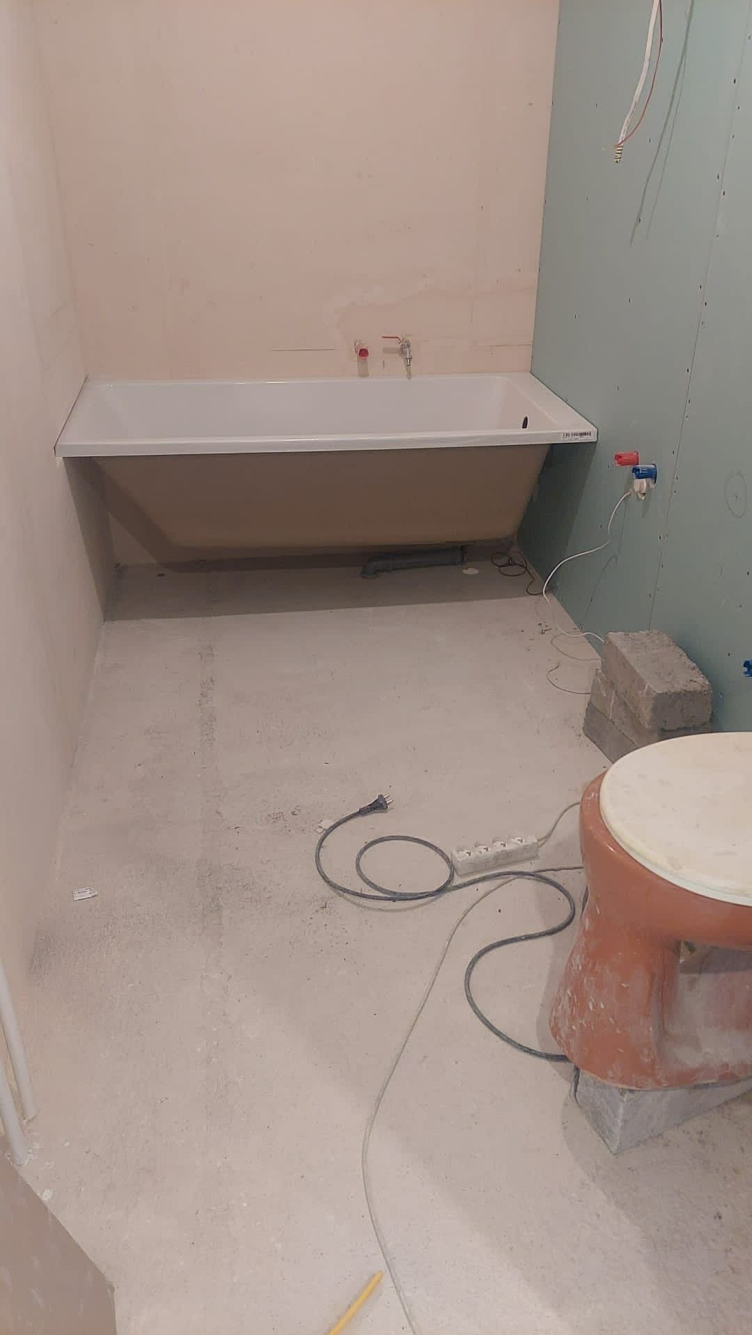 Beżowa wanna osadzona w surowej łazience z niedokończonymi ścianami i podłogą, widoczne przewody elektryczne, fragment toalety i cegły.
