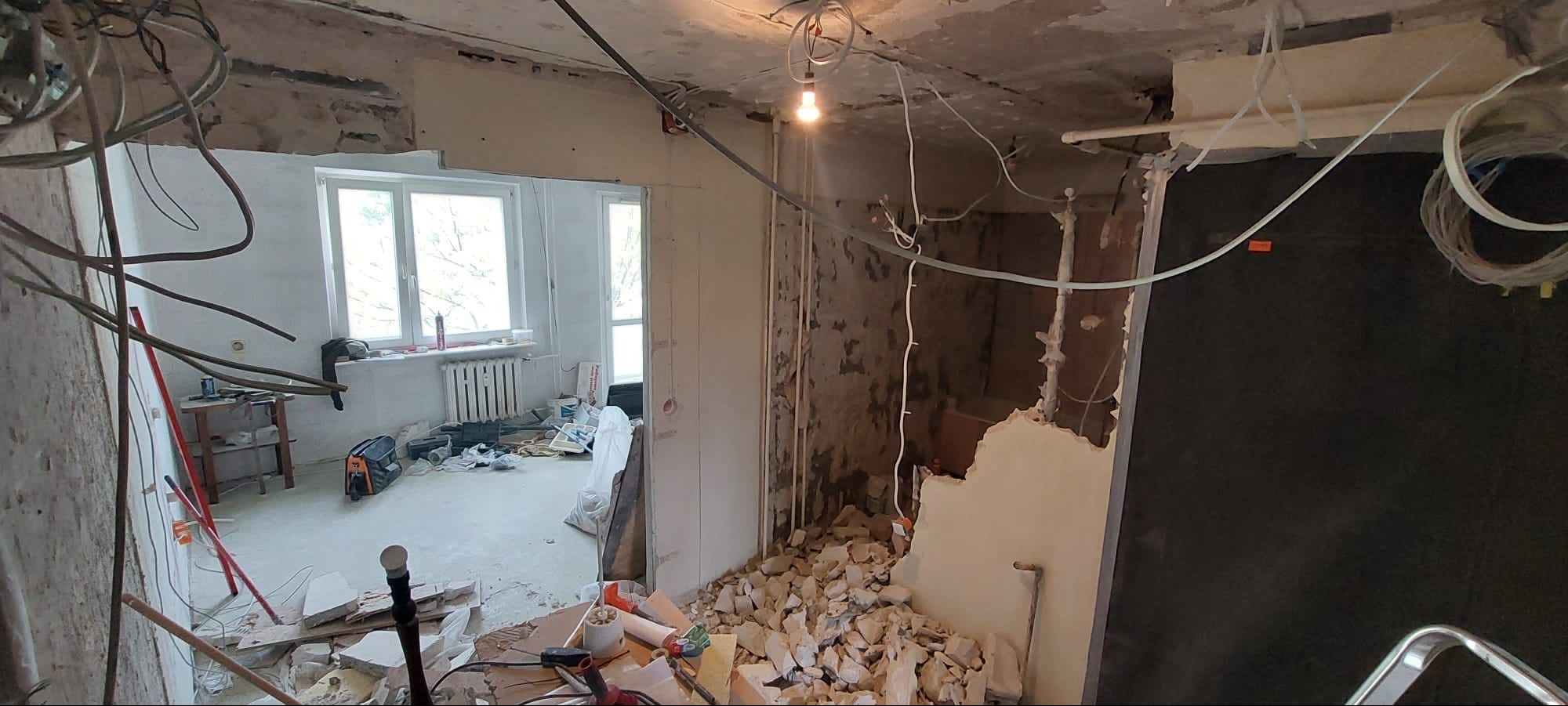 Demontaż ściany działowej w trakcie remontu mieszkania, widoczne gruzowisko, odsłonięte kable elektryczne i fragmenty instalacji, w tle okno z grzejnikiem.