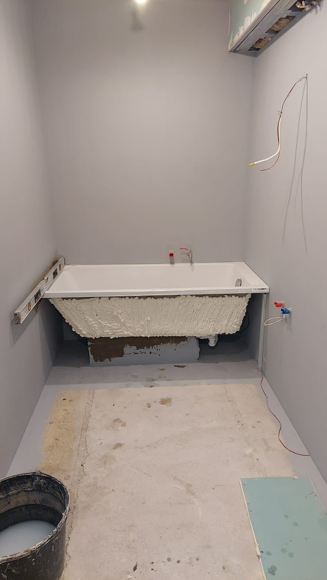 Biała wanna obłożona pianką montażową w szarej łazience w trakcie remontu, poziomica przyłożona do krawędzi, widoczne wystające przewody i rury.