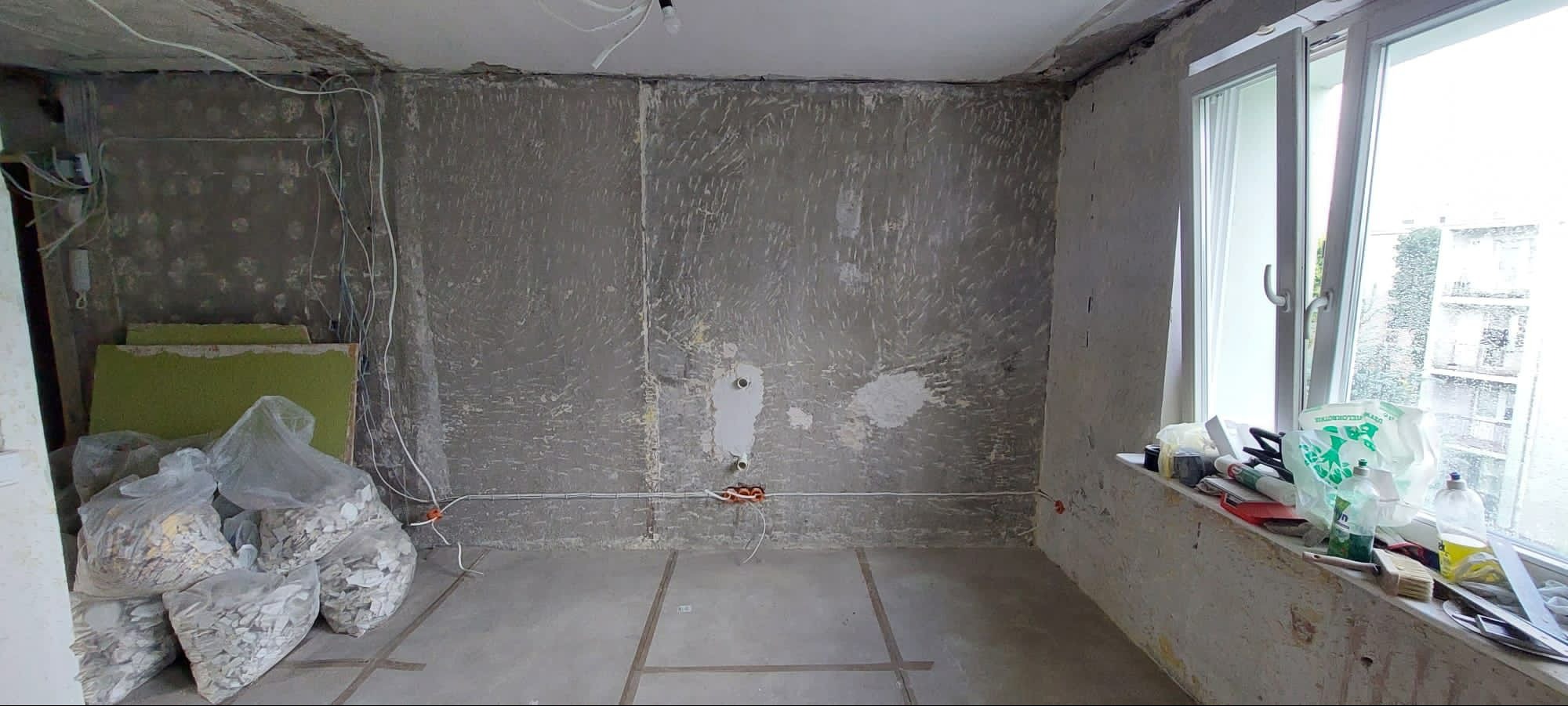 Pokój w trakcie remontu: surowe ściany z widocznymi śladami po skuciu, odsłonięte przewody elektryczne, worki z gruzem i narzędzia na parapecie okiennym.