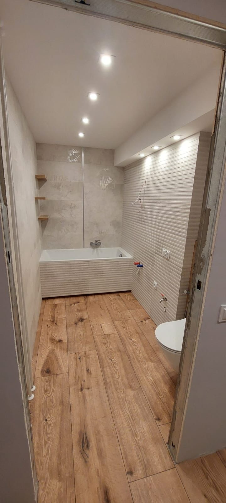 Nowoczesna łazienka z drewnopodobną podłogą, białą wanną obudowaną płytkami i ścianą z dekoracyjną fakturą. Widoczne oświetlenie punktowe i półki z jasnego drewna.