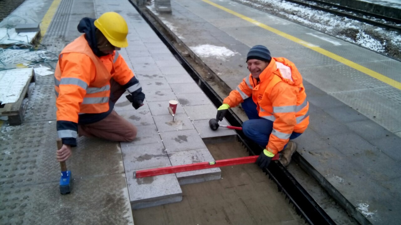 Dwóch pracowników w pomarańczowych kurtkach i kaskach układa szare płyty chodnikowe na peronie kolejowym, używając gumowego młotka, poziomicy i kielni, widoczny kubek po kawie.