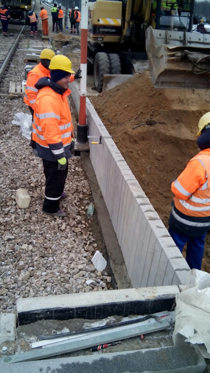 Ekipa budowlana w pomarańczowych kurtkach montuje betonowe elementy obrzeża chodnika przy torach kolejowych; w tle koparka Caterpillar.