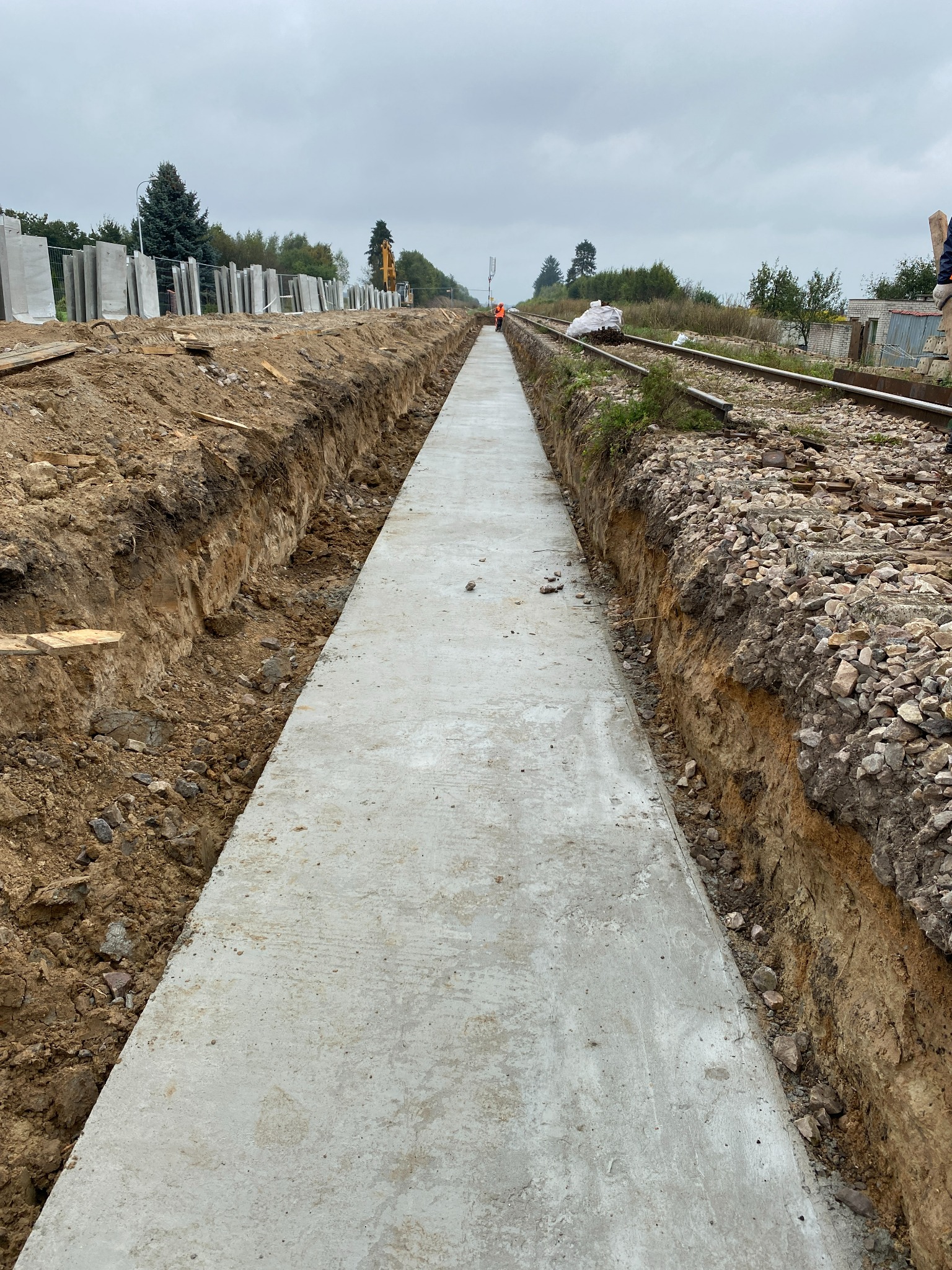 Wykop w ziemi z ułożonym betonowym chodnikiem wzdłuż torów kolejowych, w tle widoczny pracownik w pomarańczowym kombinezonie oraz koparka.