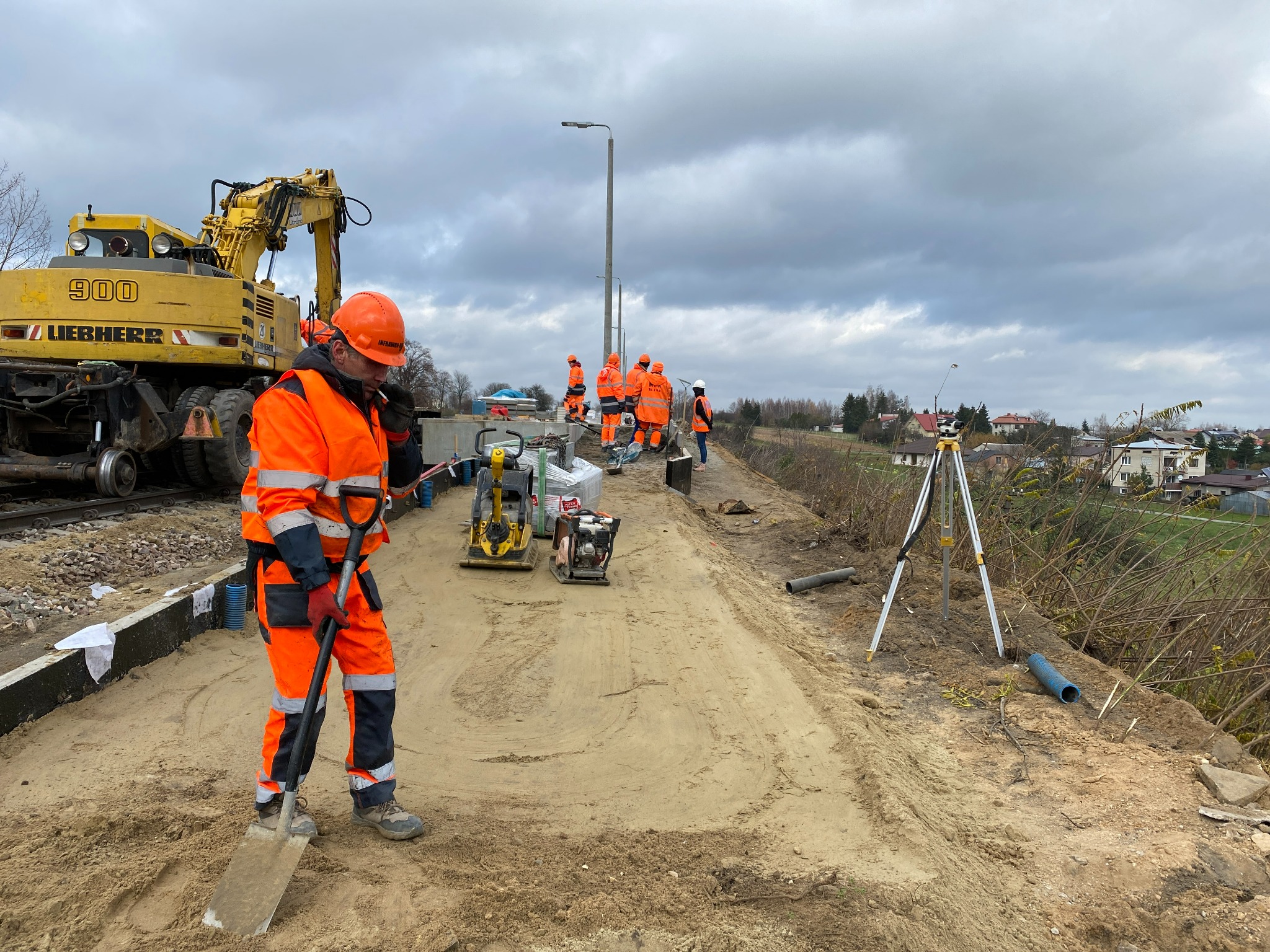 Pracownik w pomarańczowym kombinezonie z łopatą na budowie drogi, w tle ekipa, ciężki sprzęt i budynki mieszkalne.
