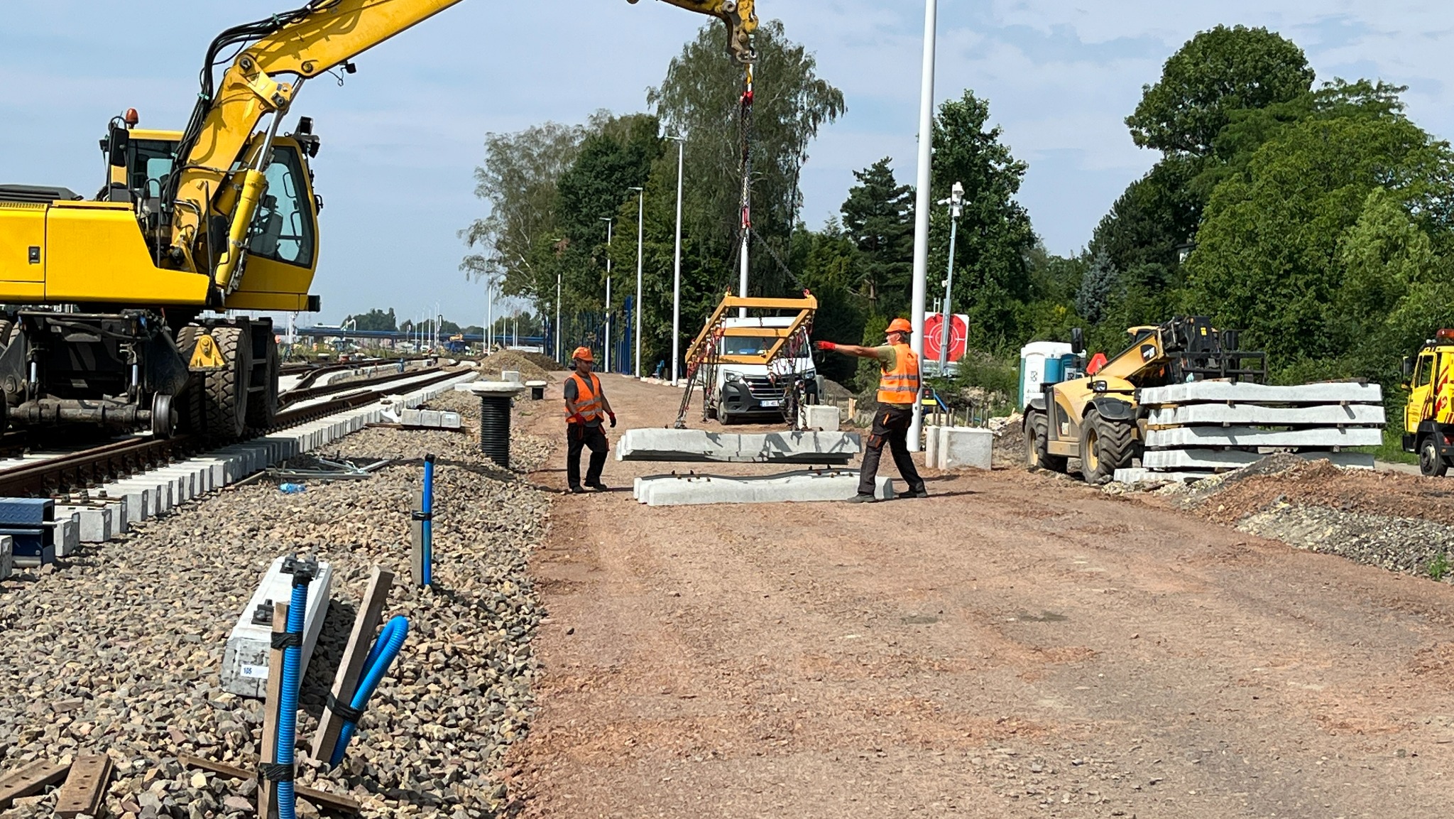 Robotnicy w pomarańczowych kamizelkach nadzorują podnoszenie betonowych elementów konstrukcyjnych dźwigiem na placu budowy torów kolejowych, widoczny ciężki sprzęt i gruz.