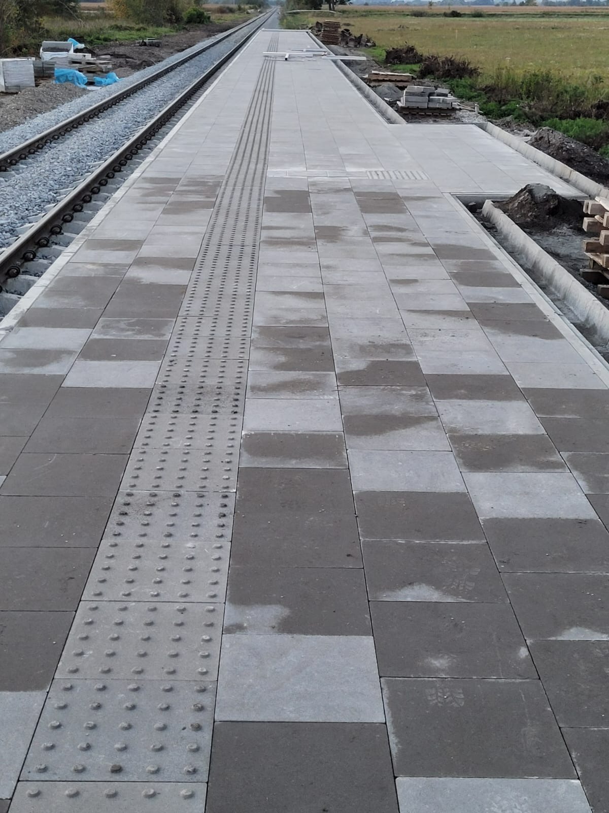 Nowa nawierzchnia peronu kolejowego z szarych, prostokątnych płyt chodnikowych, z pasem płytek z wypustkami dla osób niedowidzących, widok z perspektywy torów.