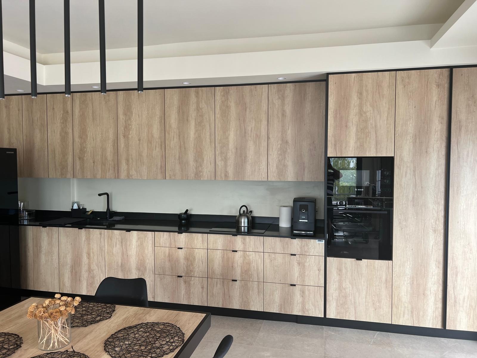 Nowoczesna kuchnia z drewnopodobnymi frontami, czarnym blatem i minimalistycznym oświetleniem. Widoczny stół z dekoracją kwiatową i czarnymi podkładkami.