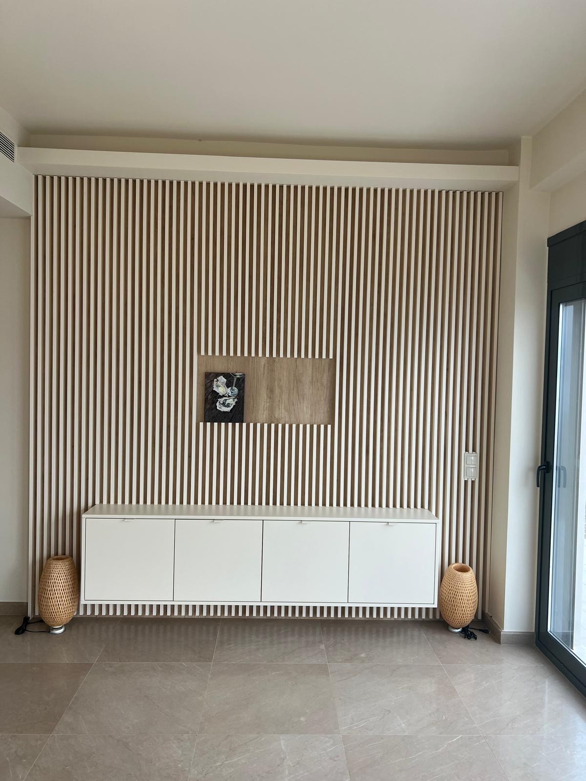 Biała szafka RTV pod ścianą z drewnianych listewek, z dekoracyjnym obrazem i lampami z rattanu po bokach. Minimalistyczny design wnętrza z jasną podłogą.