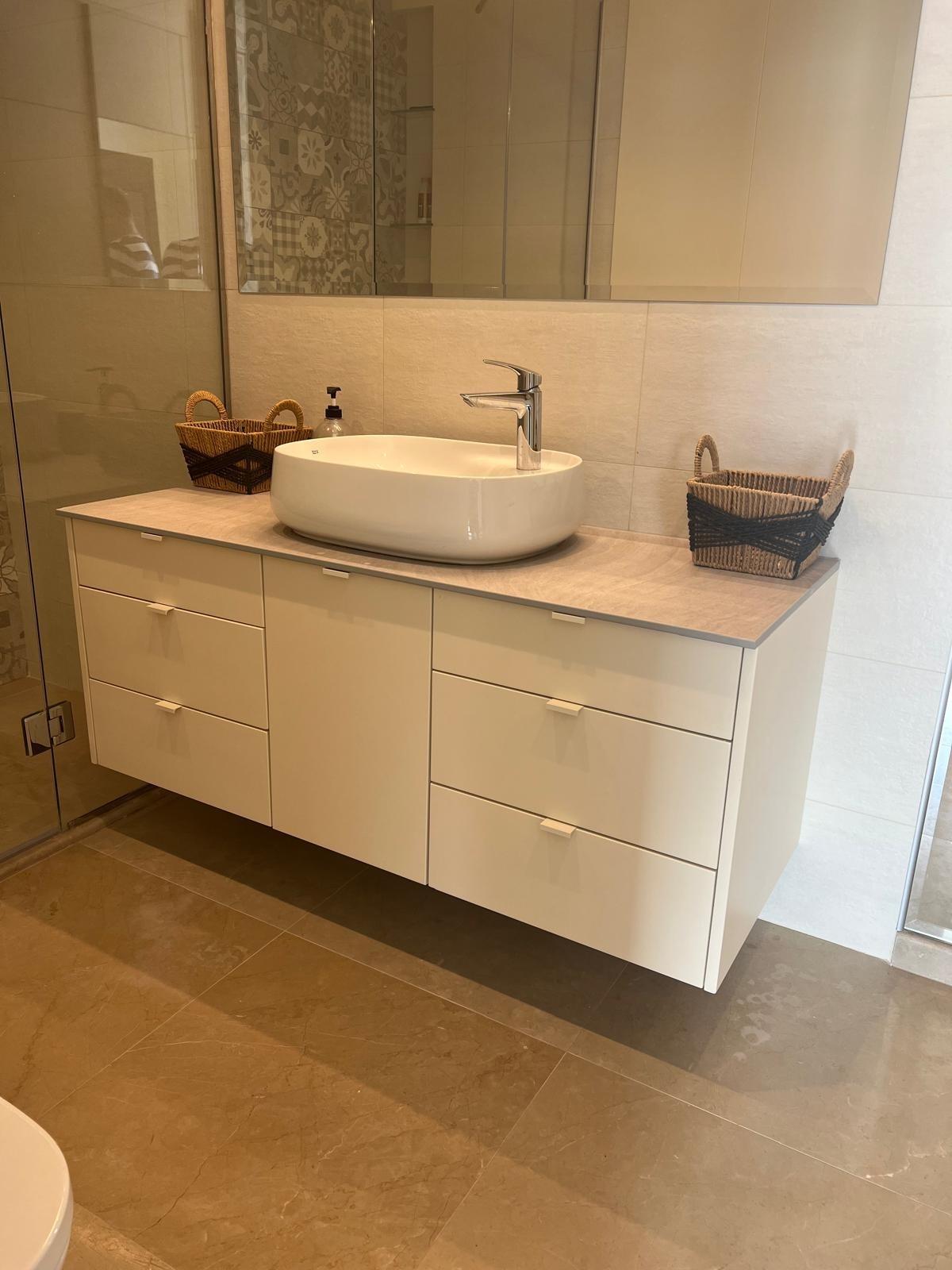 Nowoczesna łazienka z białą szafką pod umywalkę nablatową, brązowymi koszykami i beżowymi płytkami. Minimalistyczny design, jasne wnętrze.