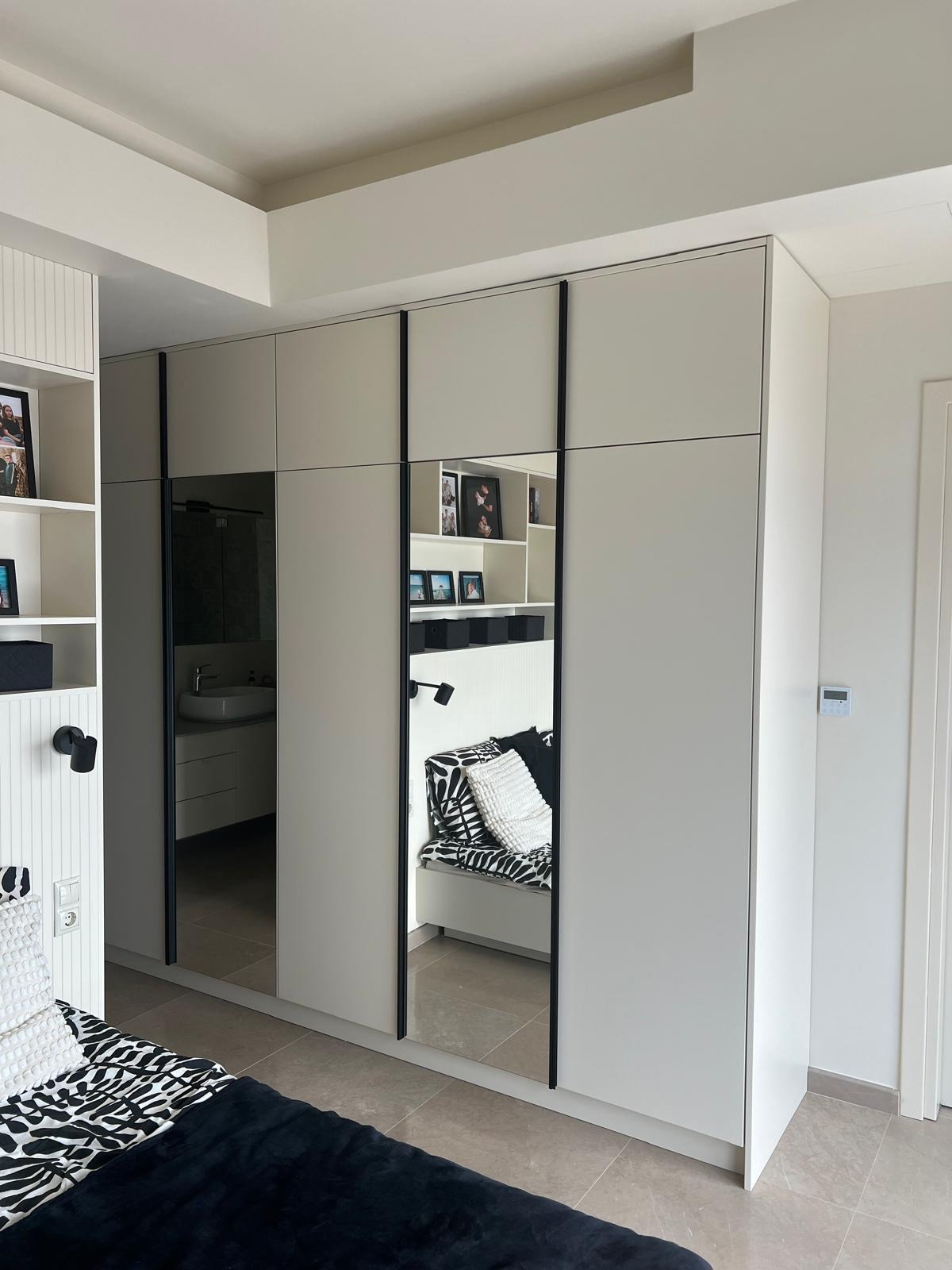 Biała szafa z lustrami w czarnych ramach, odbijająca wnętrze pokoju z półkami i siedziskiem w czarno-białe wzory. Widoczny fragment łóżka z podobnym deseniem.