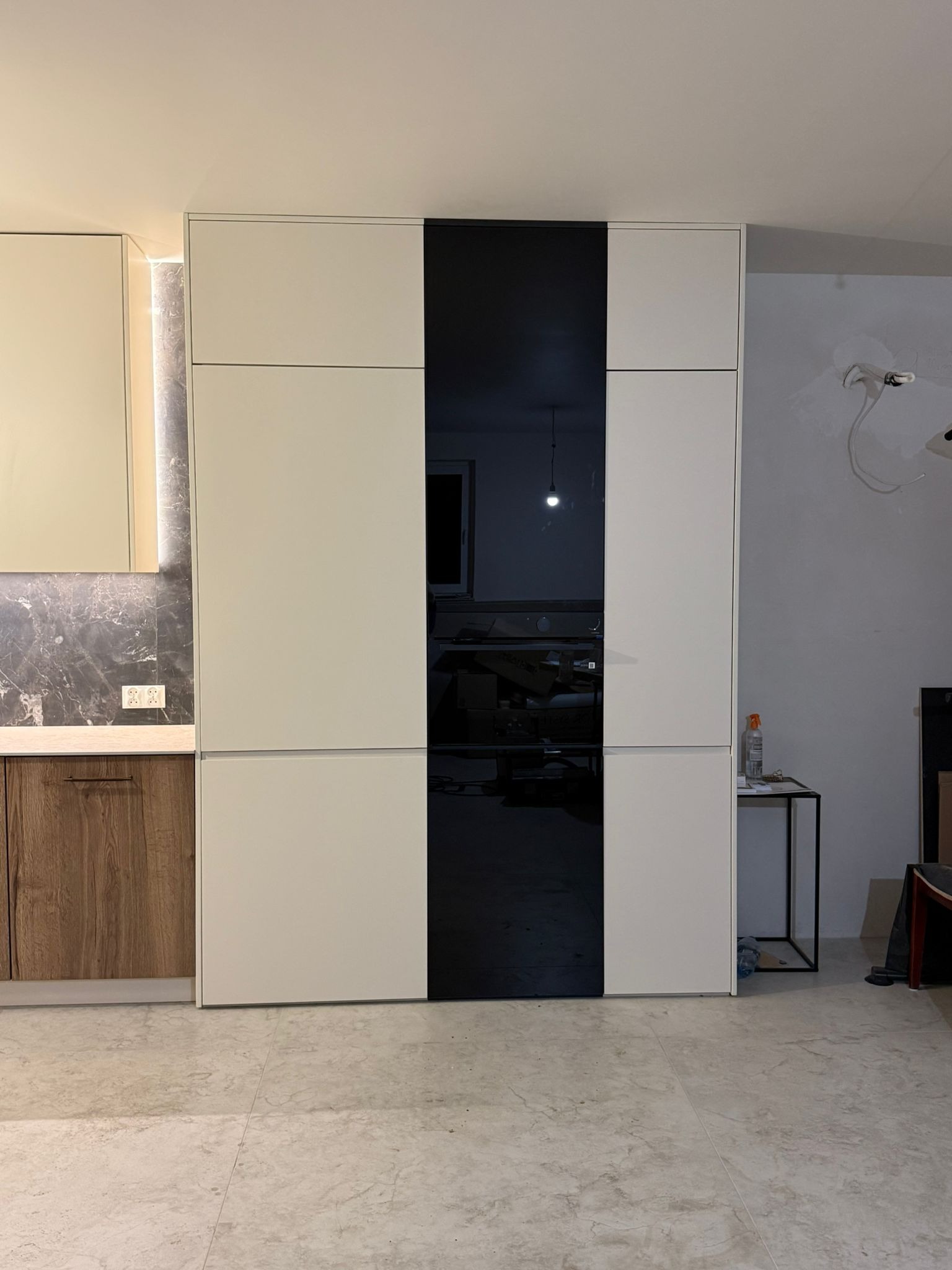 Nowoczesna zabudowa kuchenna z białymi frontami i czarnym, szklanym panelem. Widoczny fragment blatu i marmurowej ściany. Minimalistyczny design.