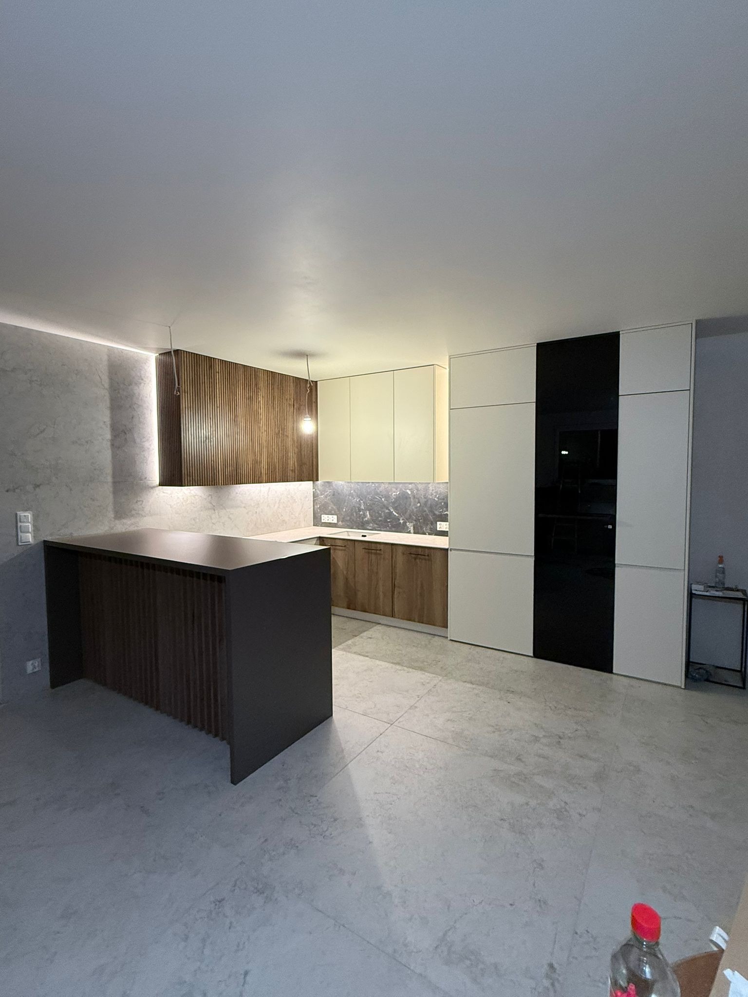 Nowoczesna kuchnia z wyspą i frontami z lameli, białe szafki z czarnym akcentem, marmurowy blat i podłoga. Minimalistyczne oświetlenie.