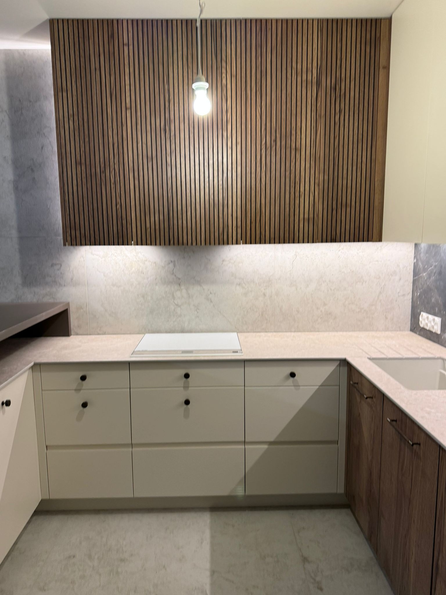 Nowoczesna kuchnia z drewnianymi lamelami na ścianie, beżowymi szafkami z czarnymi gałkami i jasnym blatem. Minimalistyczny design z wiszącą lampą.