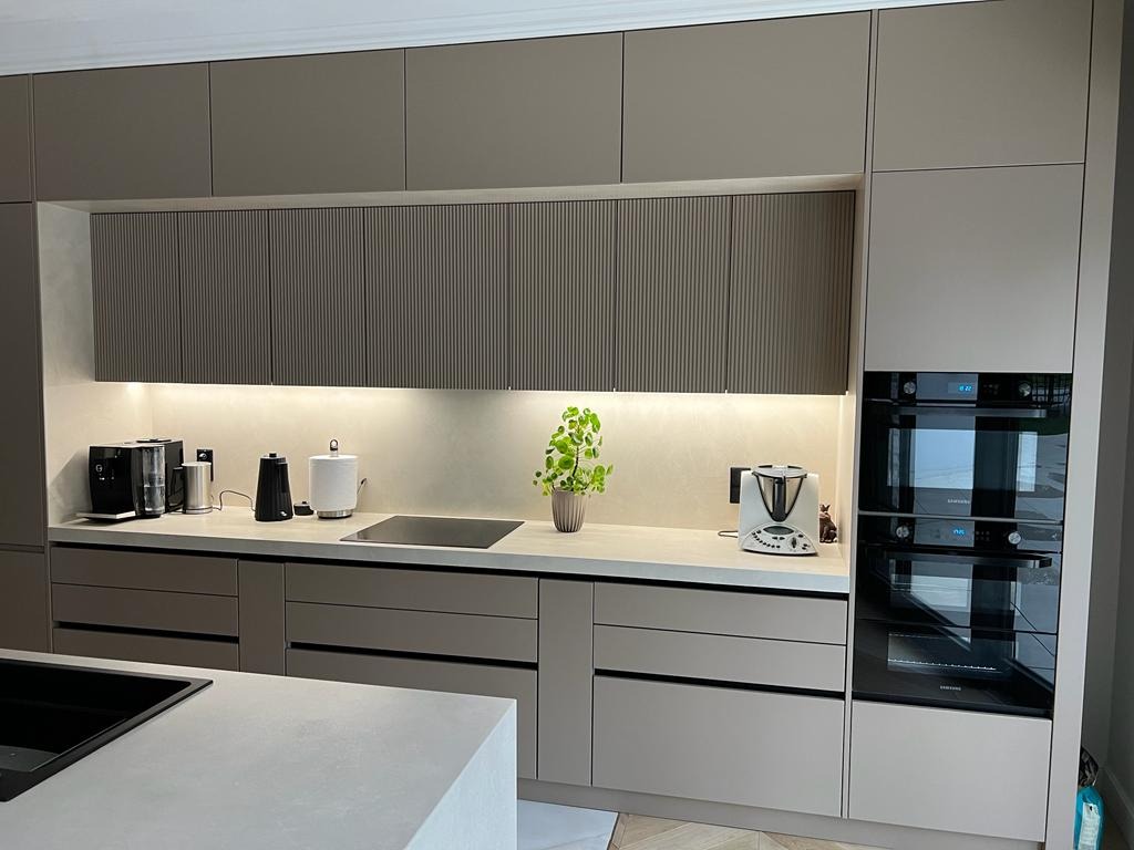 Nowoczesna kuchnia w odcieniach beżu z frezowanymi frontami szafek górnych, oświetleniem LED podszafkowym i sprzętem AGD w zabudowie. Minimalistyczny design.