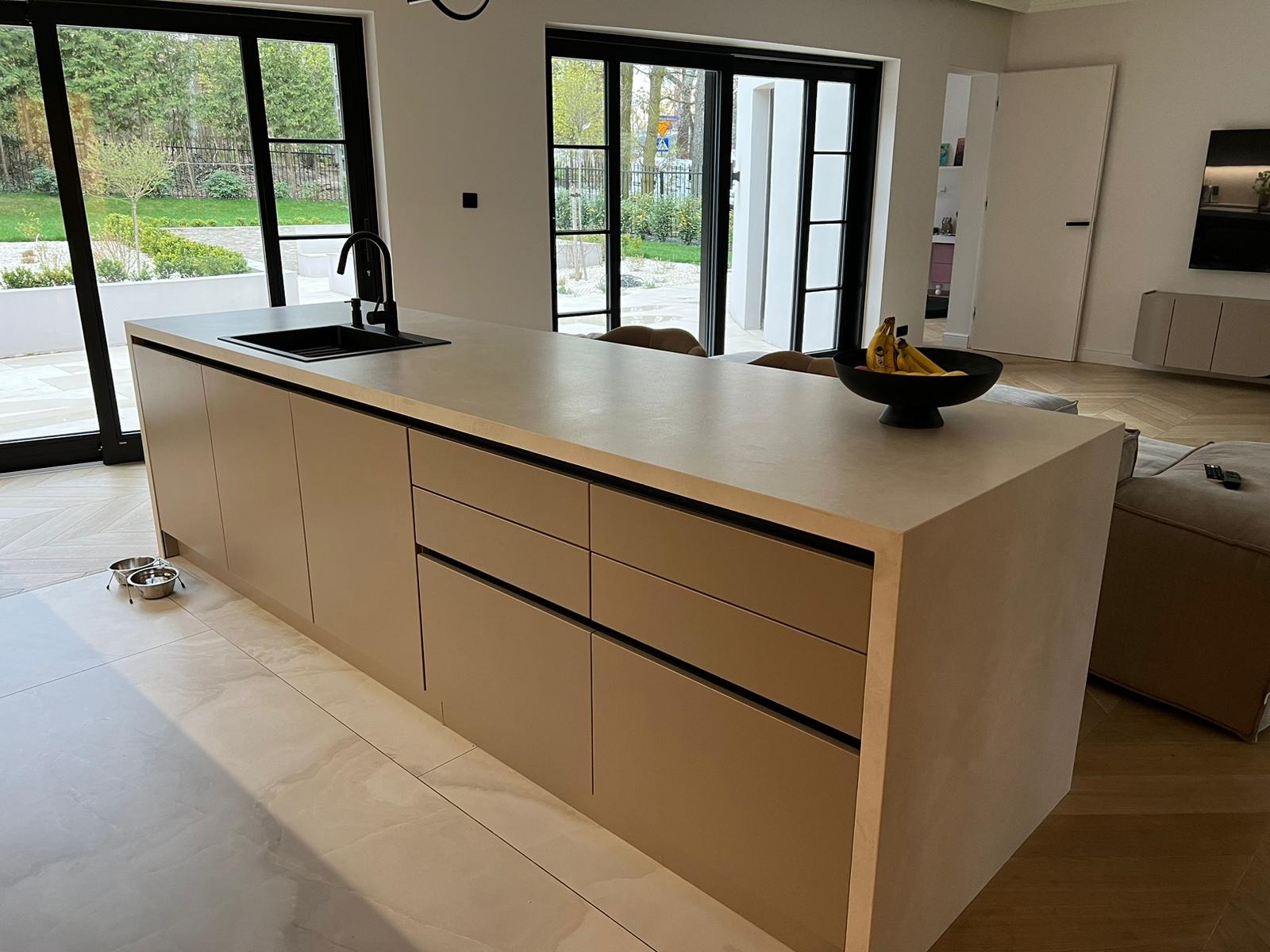 Beżowa wyspa kuchenna z czarnym zlewem i baterią, minimalistyczny design. Na blacie czarna misa z bananami. W tle duże okna z widokiem na zieleń.