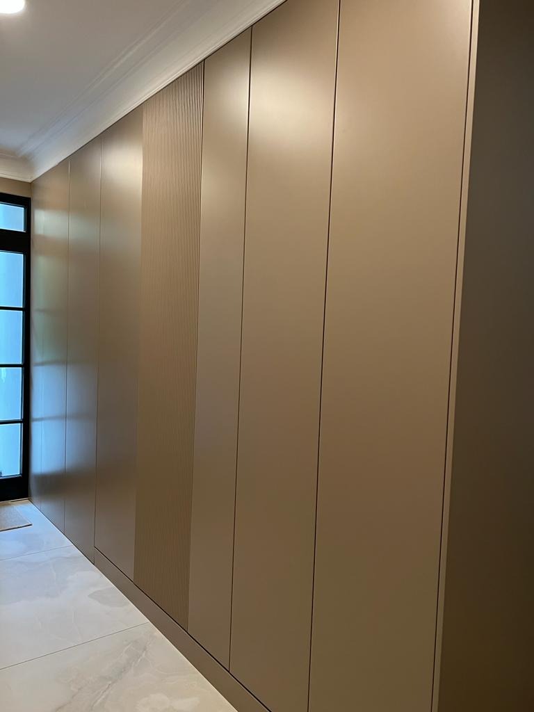 Długa, beżowa szafa wnękowa z pionowymi żłobieniami na jednej z sekcji, dopasowana do wnętrza z marmurową podłogą i listwami przysufitowymi.