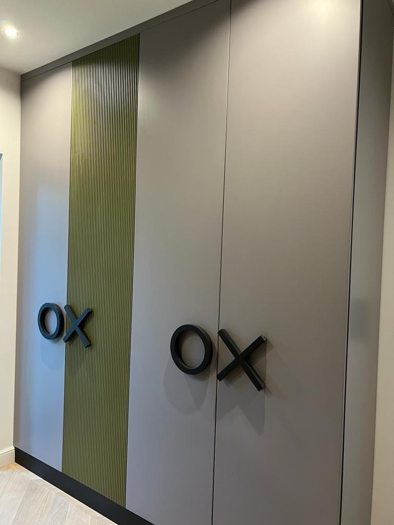 Szara szafa na wymiar z zielonym panelem i uchwytami w kształcie 'OX', tworzącymi grę kółko i krzyżyk. Nowoczesny design, minimalistyczny styl.