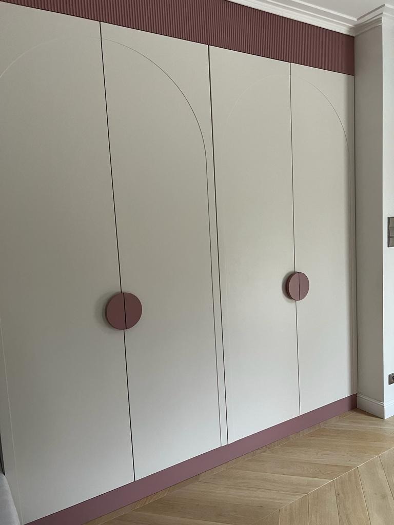 Biała szafa na wymiar z dekoracyjnymi, różowymi akcentami i okrągłymi uchwytami, wykonana w Gidlach. Minimalistyczny design z łukowym frezem na frontach.