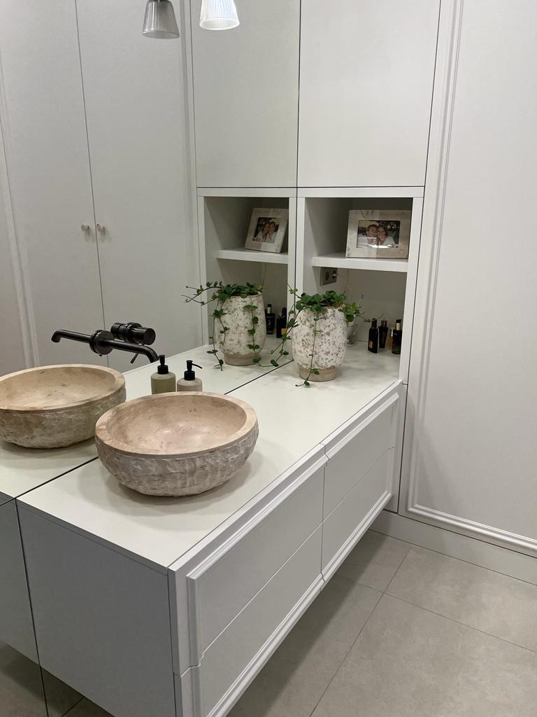 Biała łazienka z kamiennymi umywalkami i szafkami z frezowanymi frontami. Nad blatem wnęka z półkami i dekoracjami. Minimalistyczny design, jasne oświetlenie.