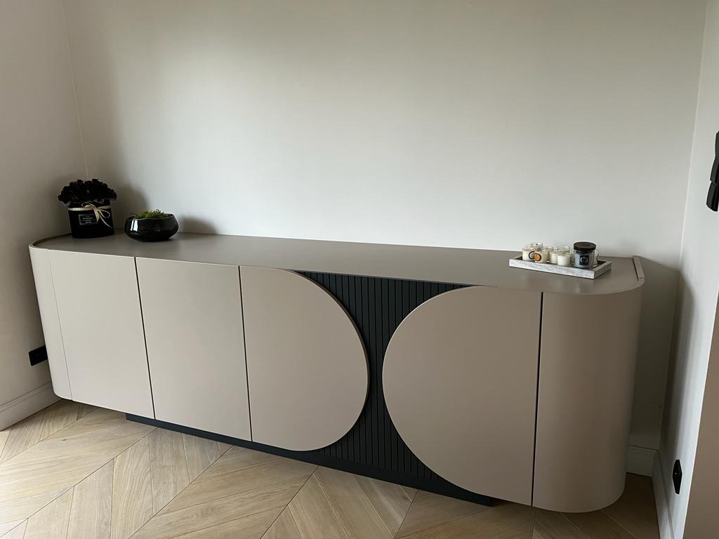 Nowoczesna komoda z zaokrąglonymi drzwiczkami w odcieniach beżu i czerni, ustawiona na parkiecie w jodełkę, z dekoracjami na blacie. Minimalistyczny design.