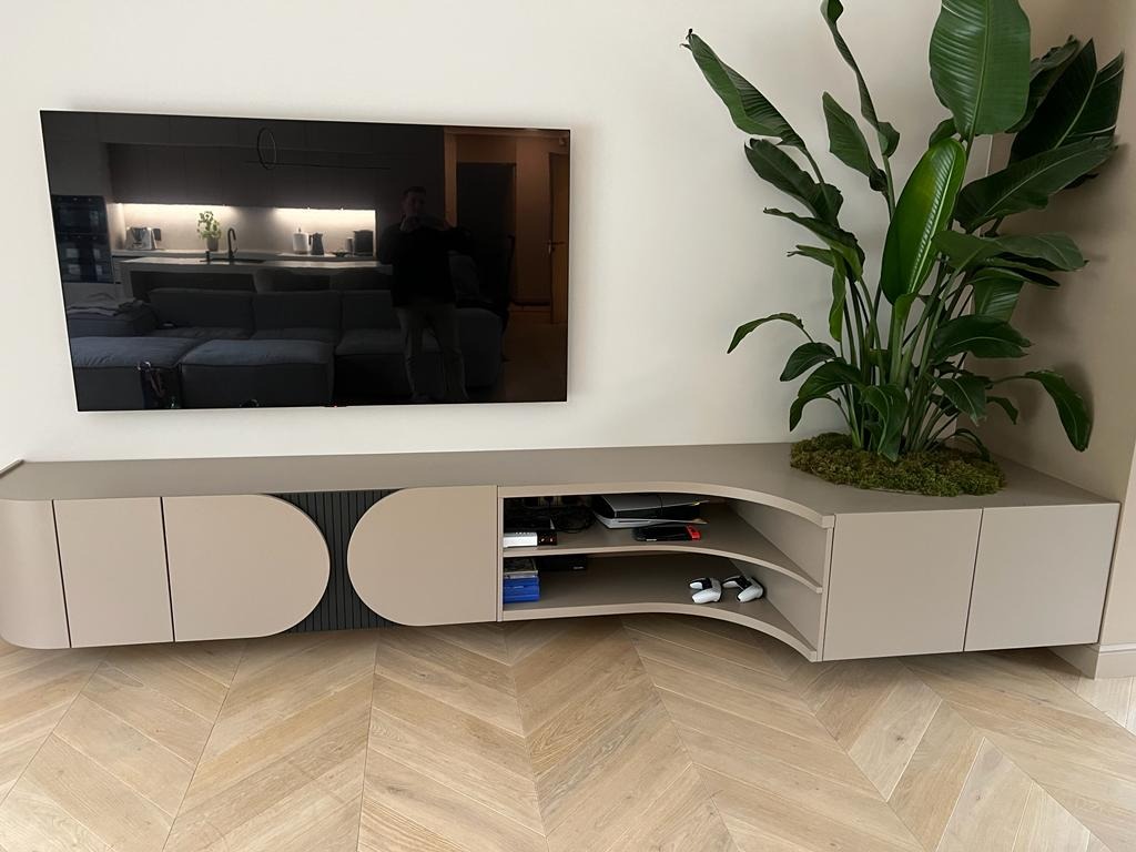 Beżowa szafka RTV z zaokrąglonymi frontami i rośliną doniczkową, na parkiecie w jodełkę. W tle widoczny telewizor i fragment kuchni.