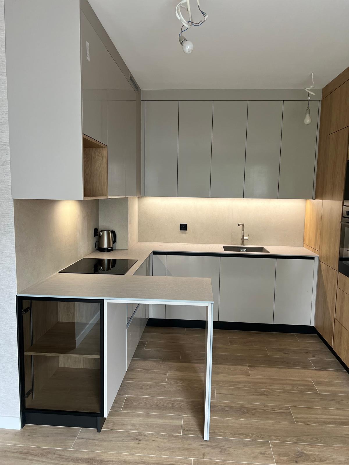 Nowoczesna kuchnia w kształcie litery U z szarymi frontami, drewnianymi akcentami i wyspą z przeszkloną szafką. Oświetlenie podszafkowe podkreśla minimalistyczny design.