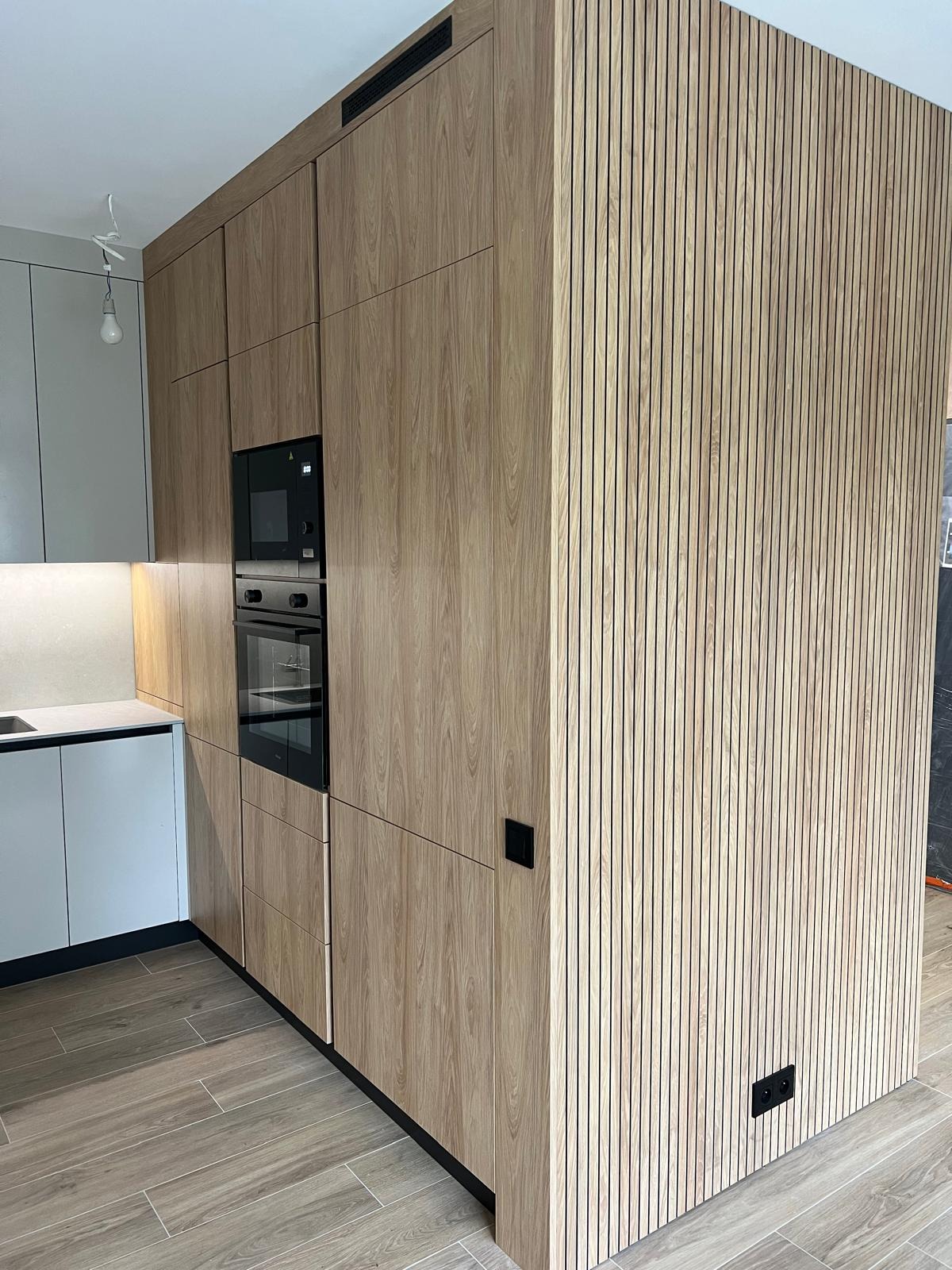 Nowoczesna zabudowa kuchenna z forniru w odcieniach drewna, z wbudowanym piekarnikiem i mikrofalówką, oraz dekoracyjną ścianką z pionowych listewek.