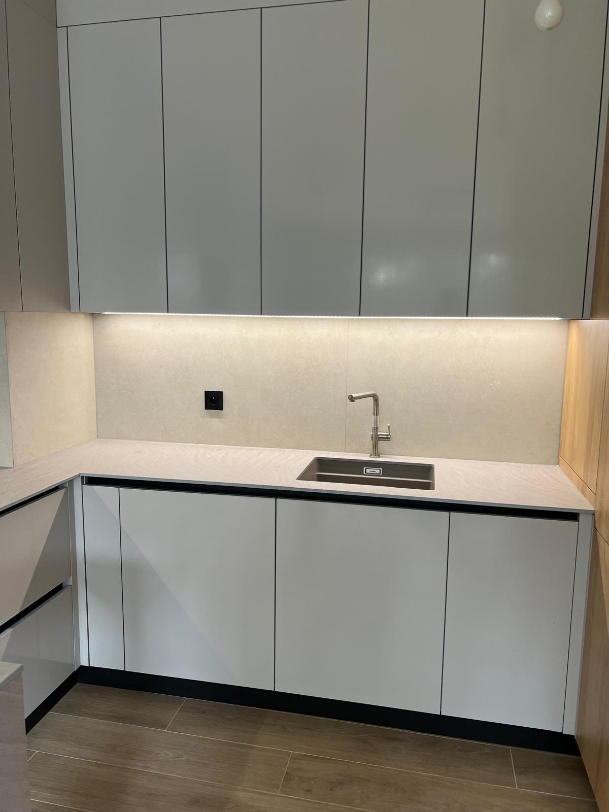 Minimalistyczna kuchnia z białymi szafkami i blatem w odcieniach beżu, oświetlona listwą LED. Uwagę przyciąga nowoczesna bateria i ciemny zlew.