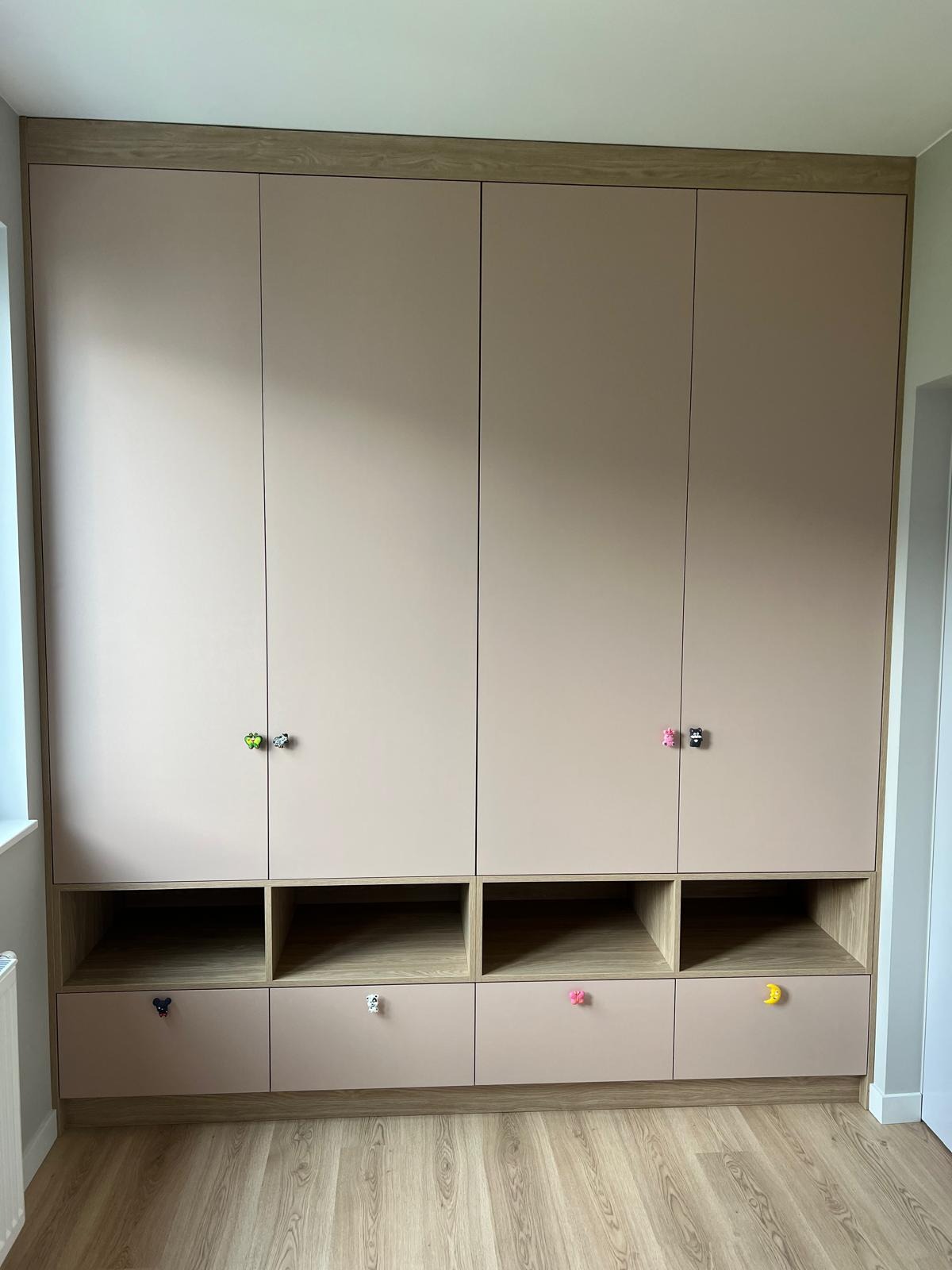 Beżowa szafa wnękowa z drewnianym wykończeniem i kolorowymi uchwytami w kształcie zwierząt, na tle jasnej podłogi i ściany.