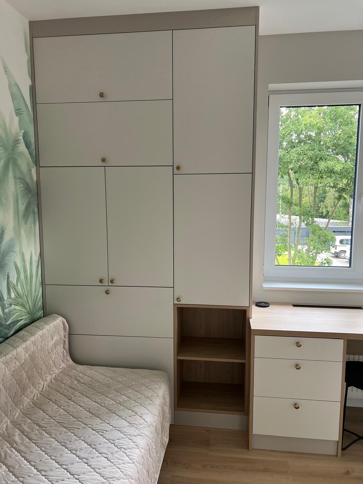 Jasna zabudowa meblowa z szafami i biurkiem, zintegrowana z tapicerowanym siedziskiem. Złote okrągłe uchwyty, drewniane półki, tapeta z motywem roślinnym.