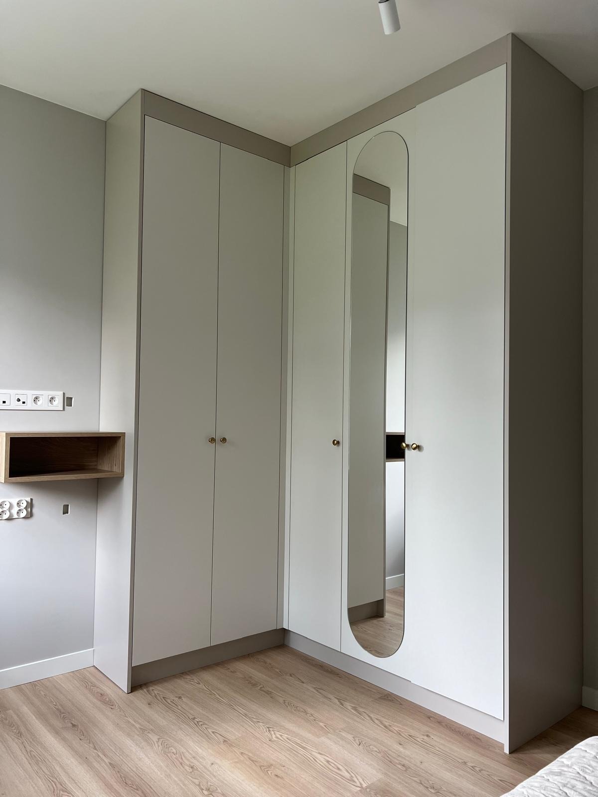 Jasnoszara szafa narożna z lustrem w kształcie łuku i złotymi gałkami, dopasowana do wnętrza pokoju z drewnianą podłogą i minimalistyczną półką.