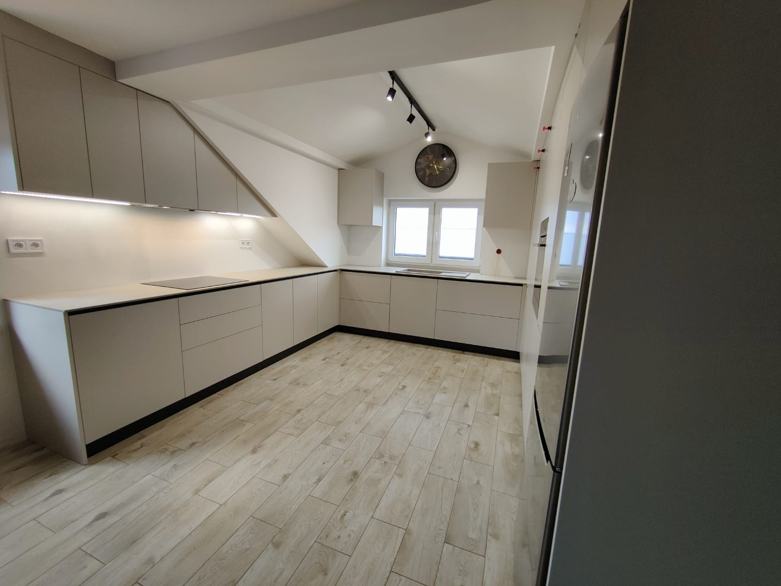 Jasna kuchnia z zabudową pod skosem, minimalistyczne fronty, czarne akcenty, drewnopodobna podłoga, oświetlenie LED podszafkowe i zegar na ścianie.