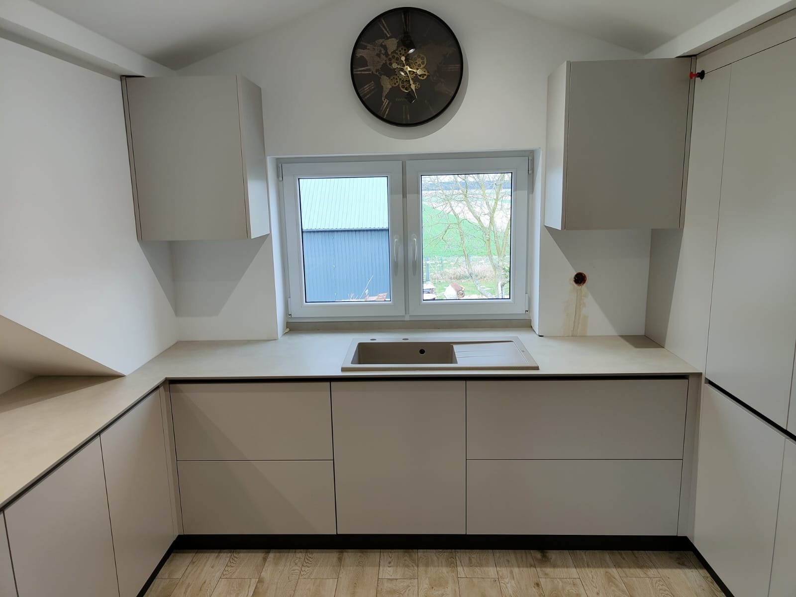 Jasna kuchnia z oknem i zegarem. Minimalistyczne szafki z ciemnymi obramowaniami, drewniana podłoga. Widok z okna na budynek gospodarczy i drzewo.