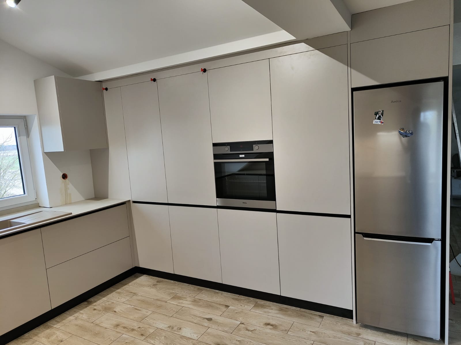Jasna kuchnia z zabudową meblową, piekarnikiem i lodówką Amica. Minimalistyczny design z drewnianą podłogą. Widoczne czerwone elementy montażowe na szafkach.
