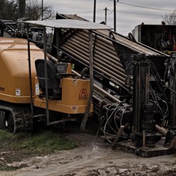HDD-WANDES SPÓŁKA Z OGRANICZONĄ ODPOWIEDZIALNOŚCIĄ - Pomarańczowa maszyna do robót ziemnych z gąsienicami i rurami w błotnistym terenie, widoczny fragment budynku i samochód w tle.