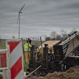HDD-WANDES SPÓŁKA Z OGRANICZONĄ ODPOWIEDZIALNOŚCIĄ - Operator w żółtej kurtce obsługuje maszynę do wiercenia horyzontalnego na tle krajobrazu z turbiną wiatrową, widoczne zabezpieczenia budowy.
