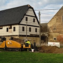 HDD-WANDES SPÓŁKA Z OGRANICZONĄ ODPOWIEDZIALNOŚCIĄ - Żółta maszyna do robót ziemnych z operatorem w kamizelce odblaskowej na tle starych budynków gospodarczych i sterty ziemi.