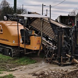 HDD-WANDES SPÓŁKA Z OGRANICZONĄ ODPOWIEDZIALNOŚCIĄ - Pomarańczowa maszyna do przewiertów sterowanych firmy Astec, z widocznymi rurami w koszu, stoi w błotnistym terenie na tle wiejskiej zabudowy w pochmurny dzień.