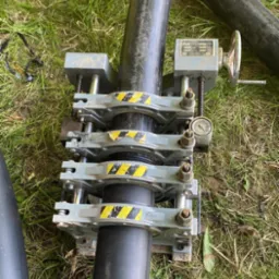 Zgrzewanie rur HDPE przy pomocy zgrzewarki doczołowej na trawie, widoczne czarne rury, złączki z żółto-czarnymi pasami ostrzegawczymi, manometr i pokrętło regulacyjne.