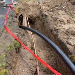 HDD-WANDES SPÓŁKA Z OGRANICZONĄ ODPOWIEDZIALNOŚCIĄ - Wykop w ziemi z widocznymi rurami: czarną, szarą i pomarańczową; zabezpieczony czerwoną taśmą ostrzegawczą, na tle zieleni i chodnika.