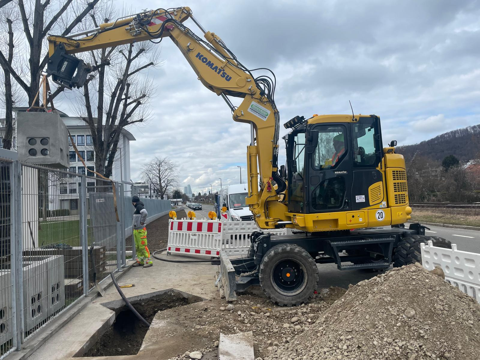 Żółta koparka Komatsu podnosi betonowy element z otworami, widoczny pracownik w odblaskowej odzieży i wykop pod rurę, w tle zabudowania miejskie.