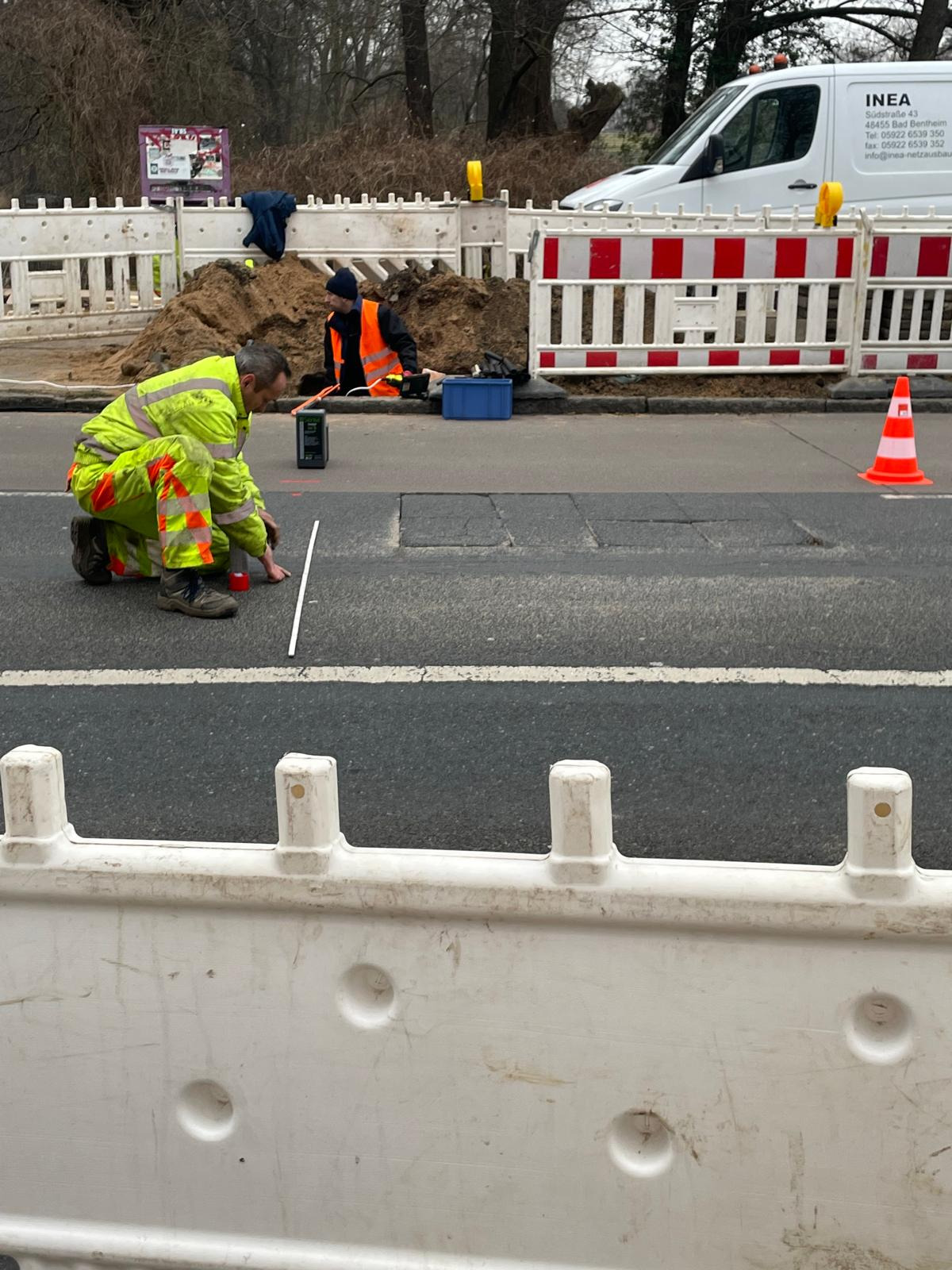 Pracownik w odblaskowym kombinezonie mierzy odległość na asfalcie przy użyciu poziomicy i dalmierza laserowego, w tle wykop zabezpieczony barierkami i drugi pracownik w kamizelce odblaskowej,...