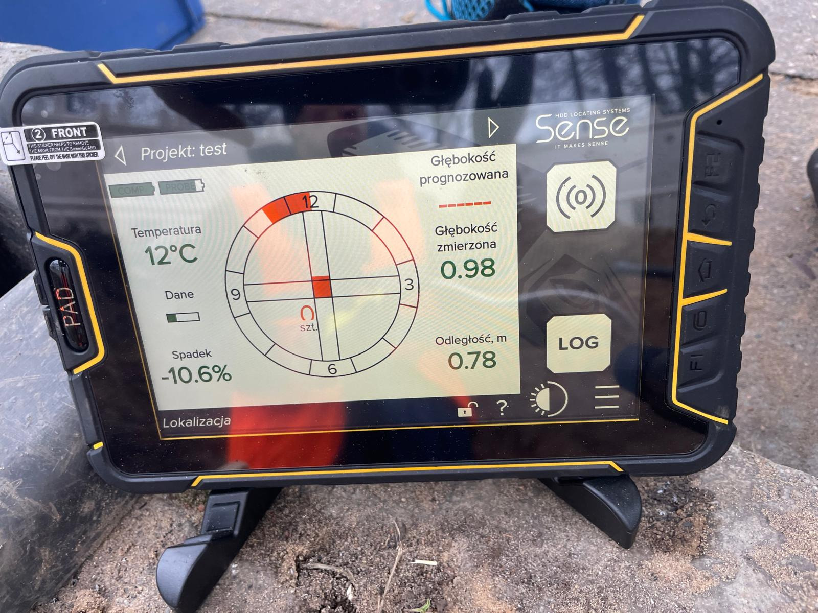 Ekran urządzenia lokalizującego HDD Sense, pokazujący dane pomiarowe głębokości, temperatury i spadku, ustawiony na szarej, kamiennej powierzchni.